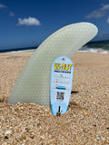 A translucent pale green surfboard fin standing upright in sand on a beach, with a blue Ez-Nut single fin screw tag inserted into the base. The background shows ocean waves and blue sky.